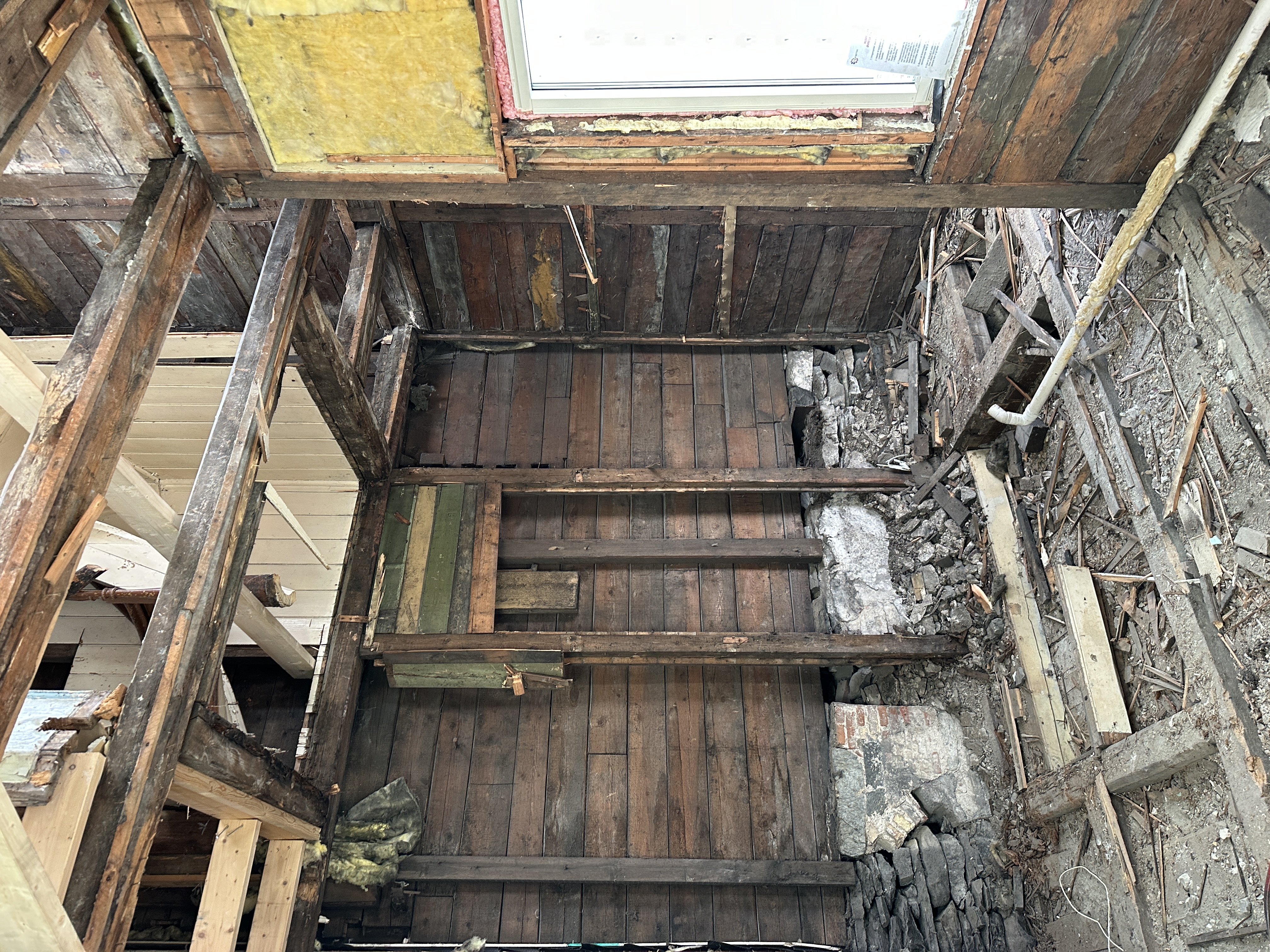 Bildet viser et pågående renoveringsarbeid i en bygning fra 1903, hvor tak og gulv er delvis revet for å avdekke den originale trekonstruksjonen. Man ser eksponerte bjelker, gamle gulvbord, isolasjon, og et nylig installert takvindu, samt bygningsrester og avfall som indikerer omfattende ombygging.