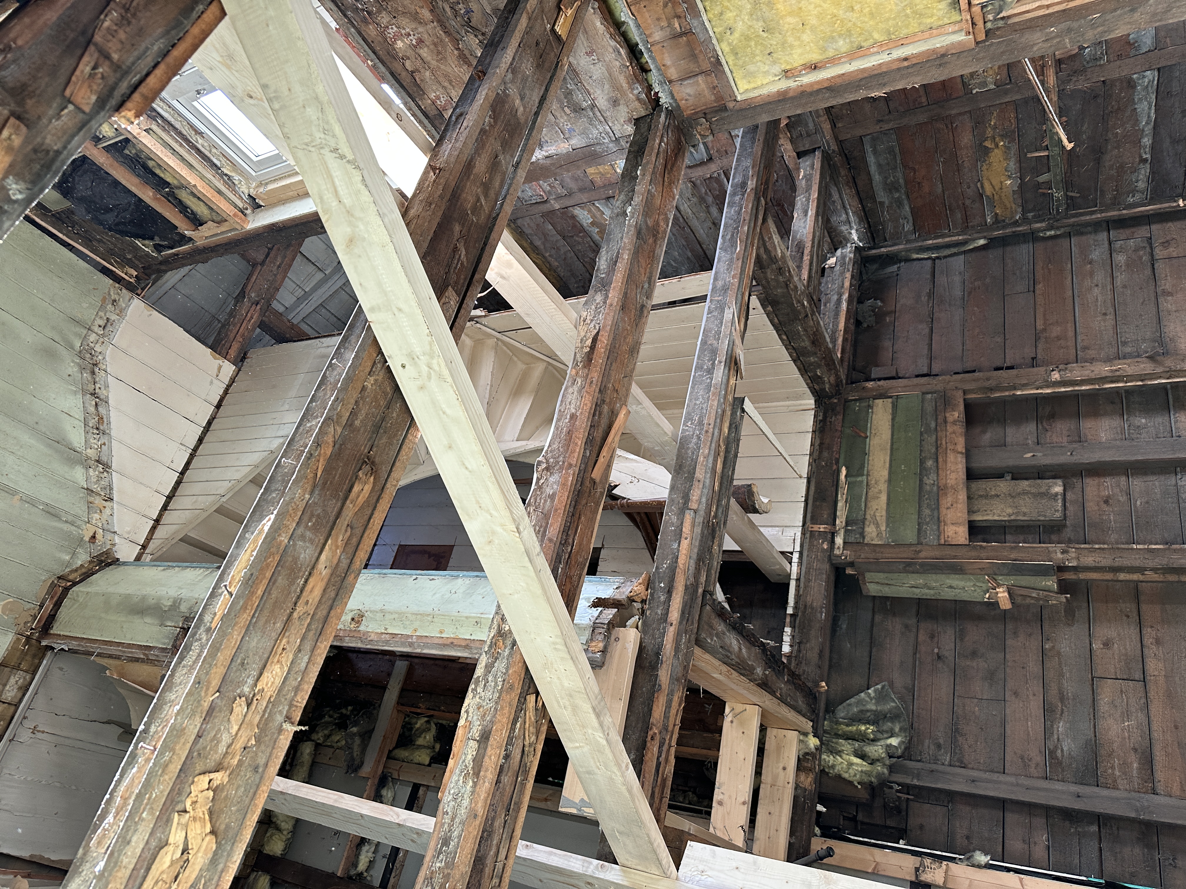 Bildet viser et pågående renoveringsarbeid i en bygning fra 1903, hvor den gamle trekonstruksjonen er eksponert og delvis forsterket med nye bjelker. Man ser rester av gamle overflater som panel og isolasjon, samt et takvindu, noe som indikerer en omfattende ombygging av loft eller takkonstruksjon.