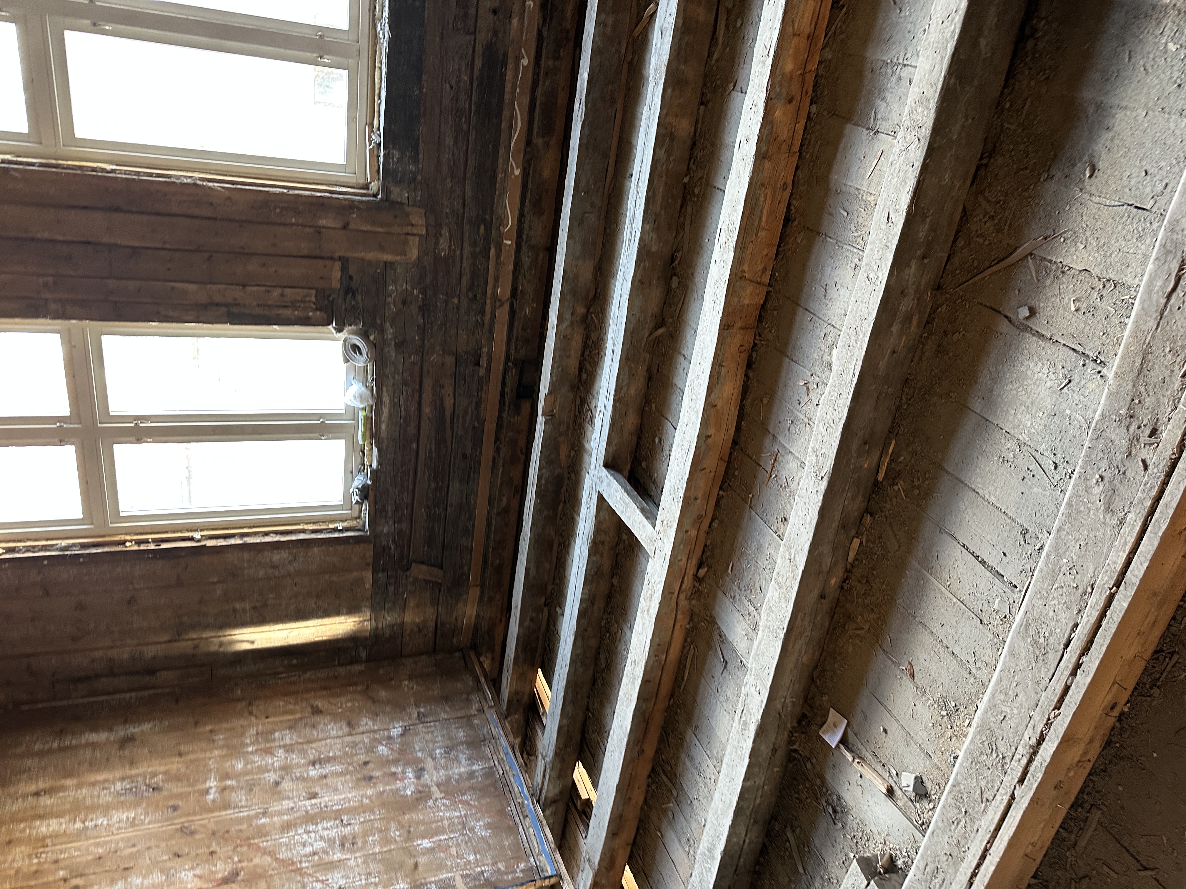 Bildet viser et rom under renovering, der vegger og tak/gulv er strippet ned til den originale trekonstruksjonen fra 1903. De eksponerte gulvbjelkene og det gamle undergulvet indikerer omfattende restaureringsarbeid.