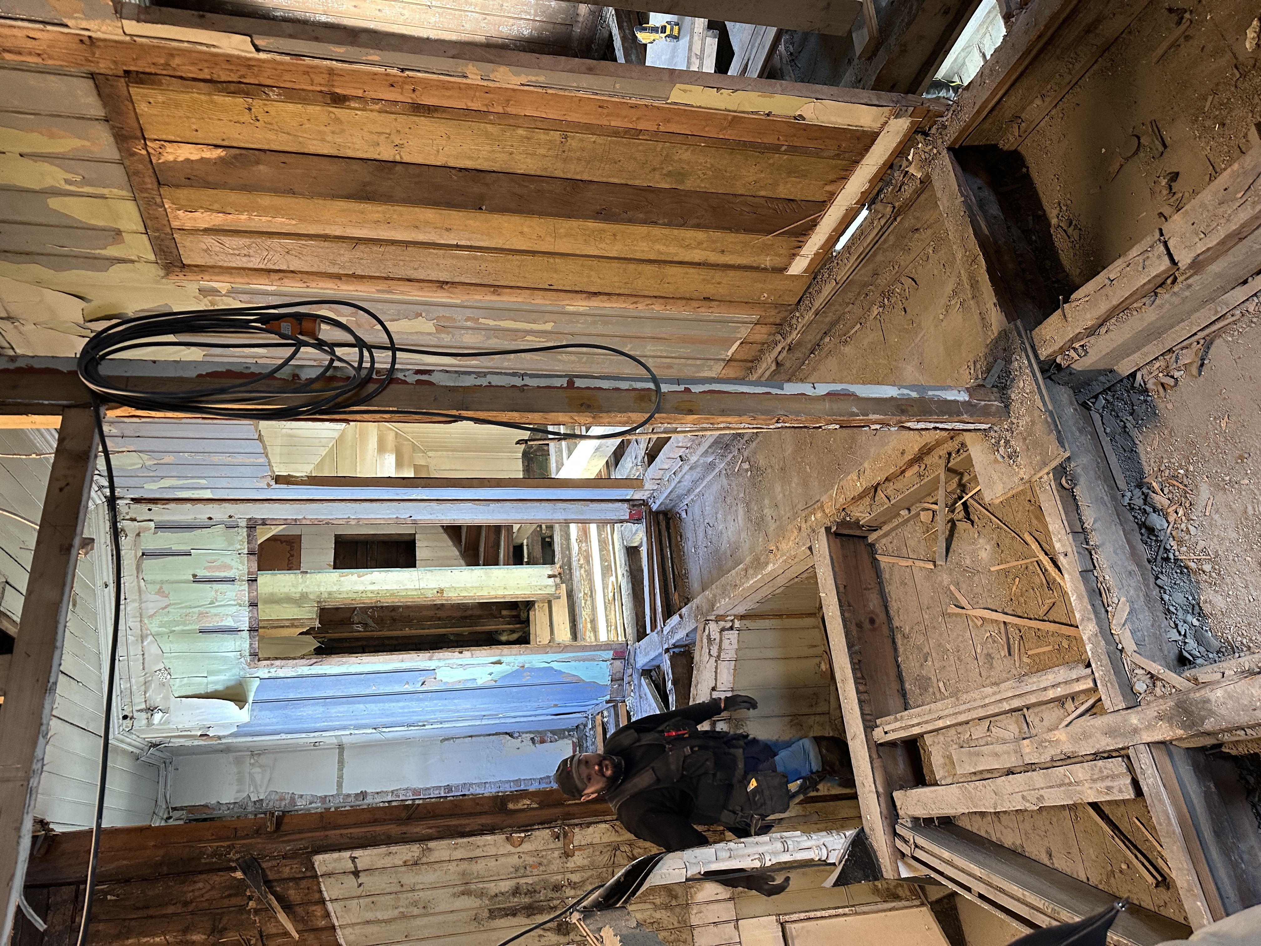 Bildet viser interiøret av en bygning fra 1903 under omfattende renovering eller rivning, hvor vegger, tak og gulv er strippet ned til den originale trekonstruksjonen. Man ser tydelig lag av gammel maling og puss, samt en arbeider som overvåker prosessen i den avkledde strukturen.
